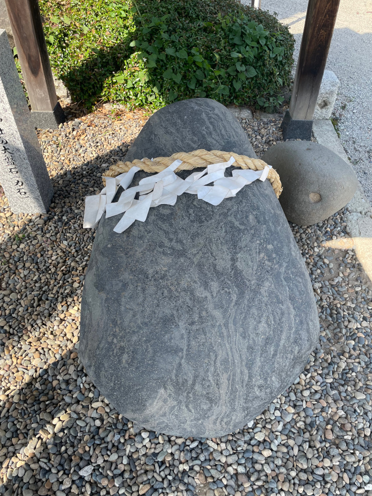 A massive stone is lying on gravel under a canopy. A shimenawa 'enclosing rope' is wrapped around the top of the stone with shide paper streamers.