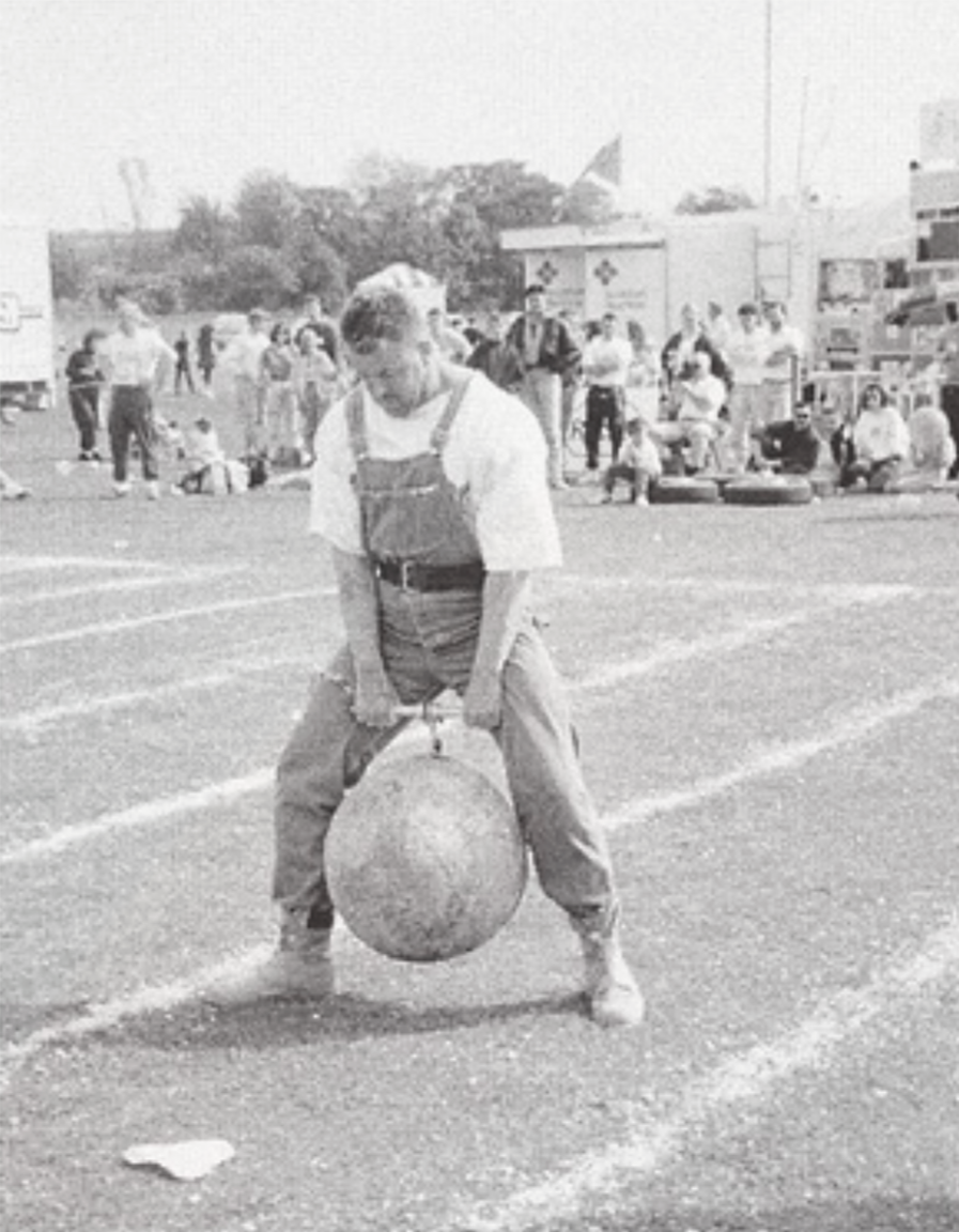 A greyscale image of a man in dungarees and a lifting belt performing a duck walk with the Dreghorn Stone