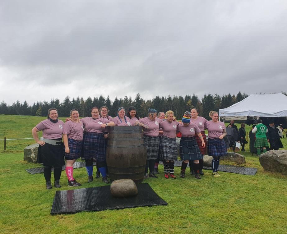 A group of women in kilts and competition T-Shirts stand around a barrel in the rain. A stone is in front of the barrel.