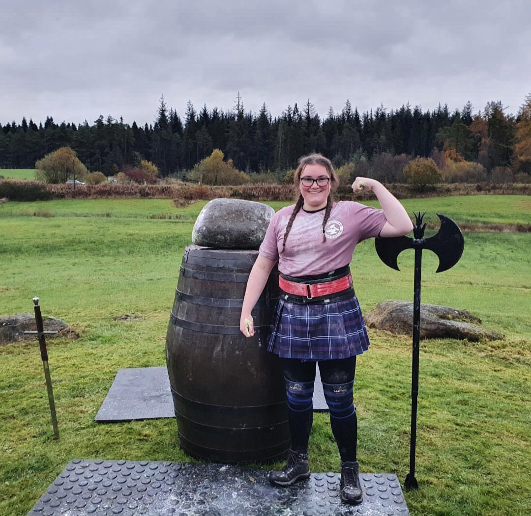 A smiling woman poses with her biceps flex in front of a barrel. The Puterach Stone sits atop the barrel.