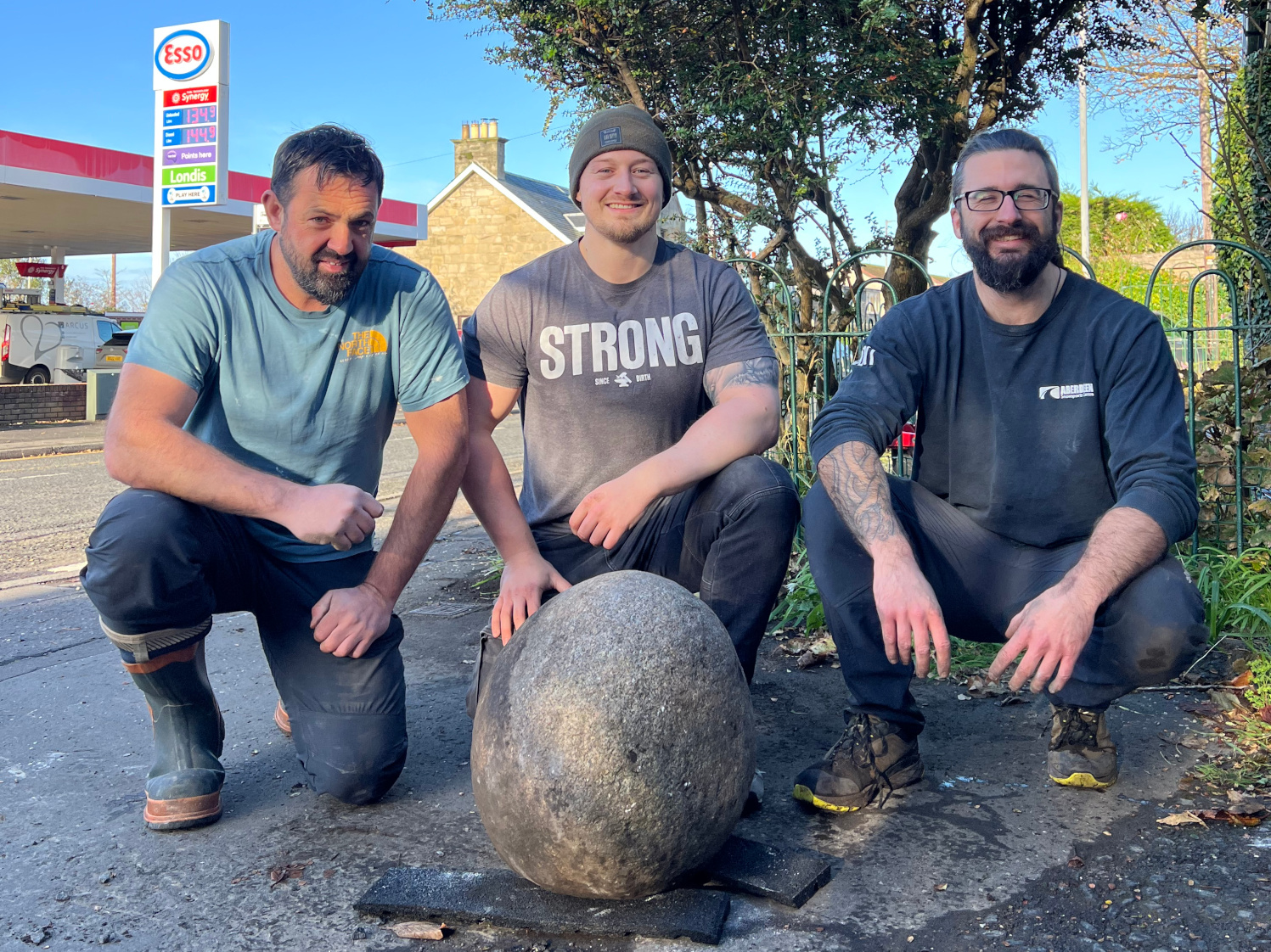 Three men crouching down for a photograph with a large stone in the forefront.