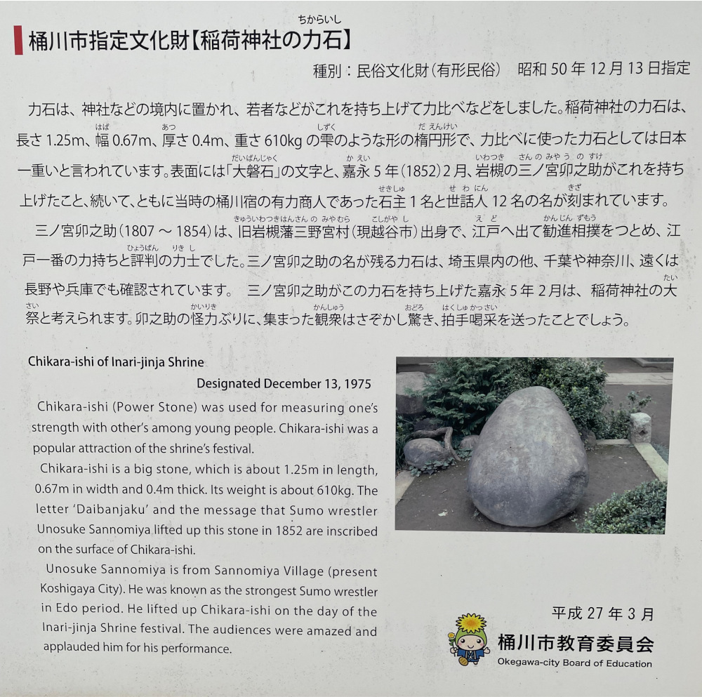 A photo of a sign placed by Okegawa City's Board of Education at the Inari Jinja in Okegawa. The sign has both Japanese and English writing with information about the 610kg stone at the shrine and Sannomiya Unosuke.