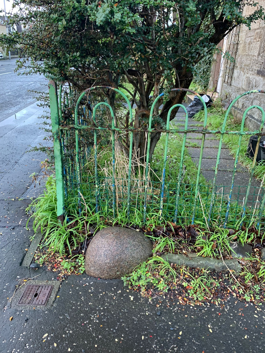 A photo of the Loans Stone buried in the pavement. It looks wedged between a thin metal fence and the pavement. About one third of the top of the stone is visible.