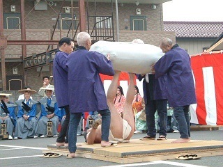 A man is lying on a small wooden platform with his legs in the air wearing a mawashi. Four other men are lifting a large, soft, pillow-like object in the shape of the massive stone to the man's feet to demonstrate how Sannomiya lifted the 610kg stone.