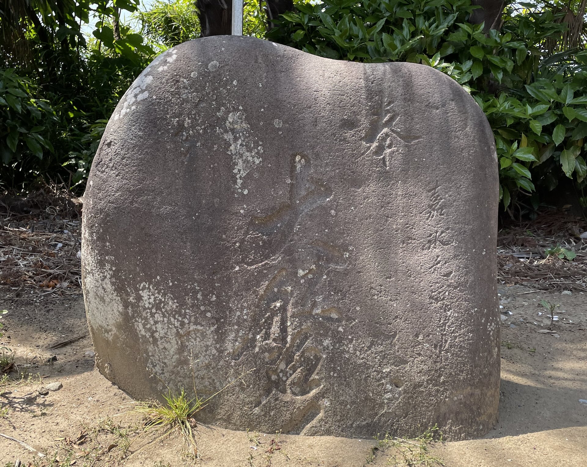 A large rounded stone with carved Japanese characters sits on bare earth in front of green bushes at Sannomiya Katori Shrine.
