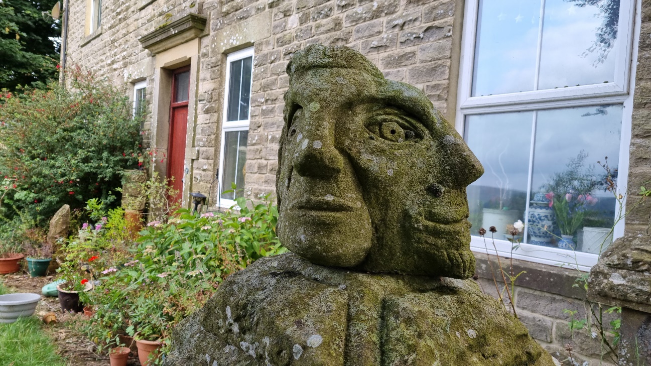 In front of a house there's a stone carving of two faces merged together on the same pedestal - the right head shares its right eye with the left head's left eye. The left head looks ahead, while the right head is at a 90 degree angle.