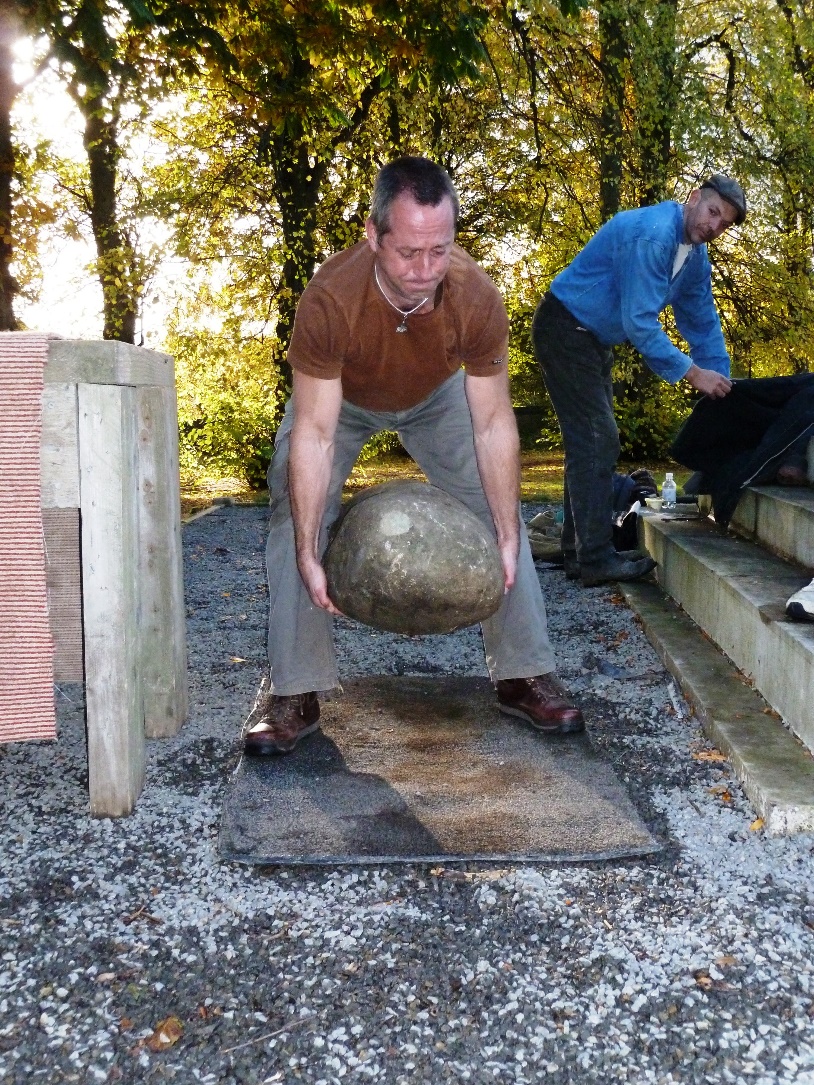 A man is bent over a large stone, gripping it from both sides. The stone is about 30cm in the air. On the top left-hand side of the stone's visible face, there's a distinctive pale circle.