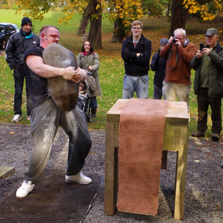 A man bearhugging a smooth, long stone. He's moving towards a waist-height wooden table.