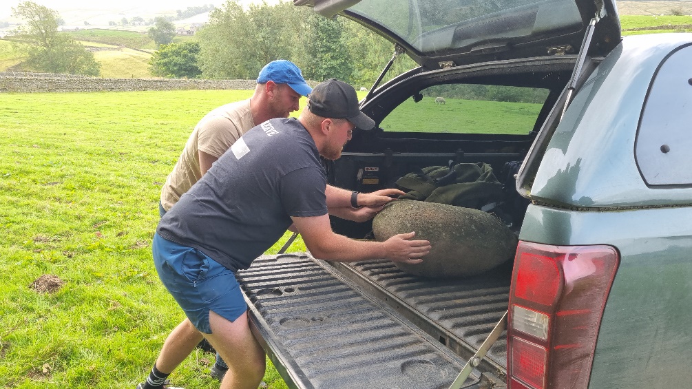 Two men push a large stone along the bed of a dark green pickup truck.