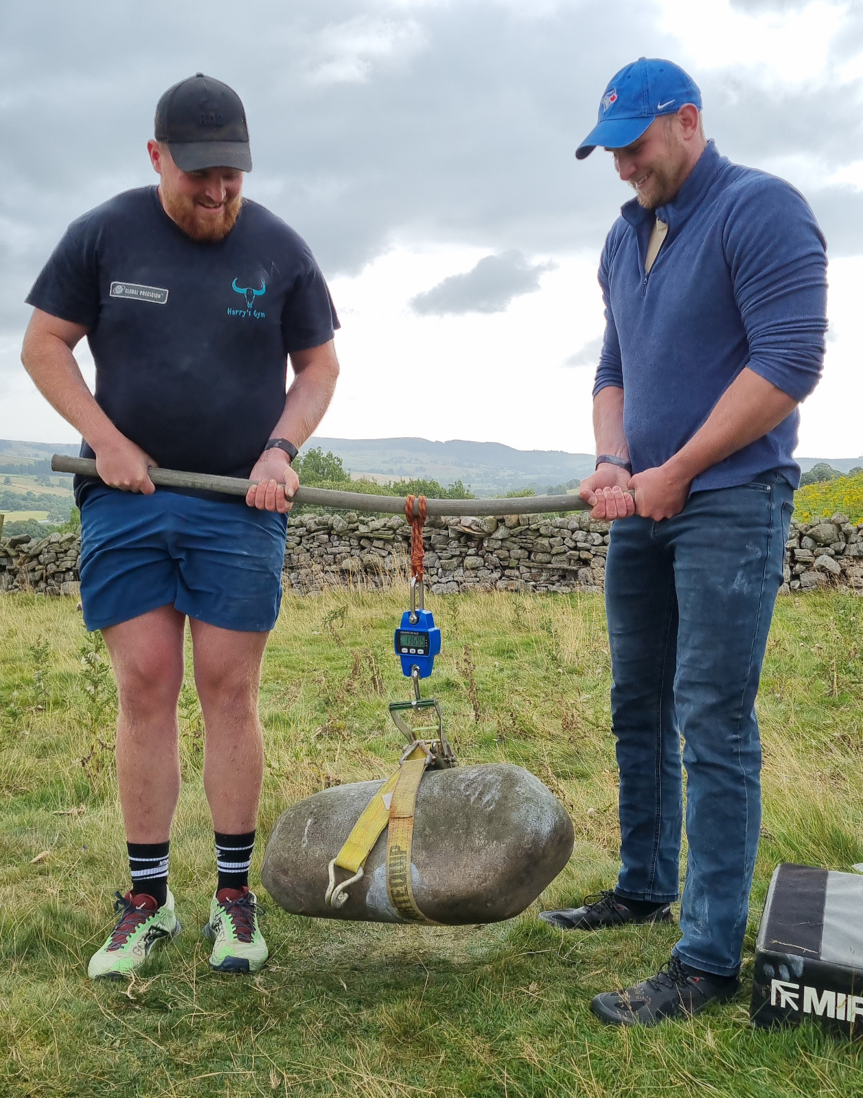 Harry and Jono stand on each end of a slightly bent metal bar. The stone dangles from the mid point of the bar wrapped in a harness attached to some scales.You can just make out the scale's screen as reading 136.95kg.