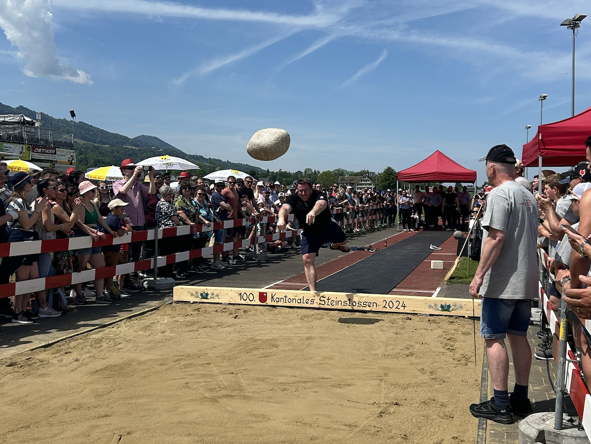 A photo of the Ybrig Stone in flight towards a sandpit. A man has just thrown the stone and is fully extended on one leg at the toe board. A crowd of people are spectating.