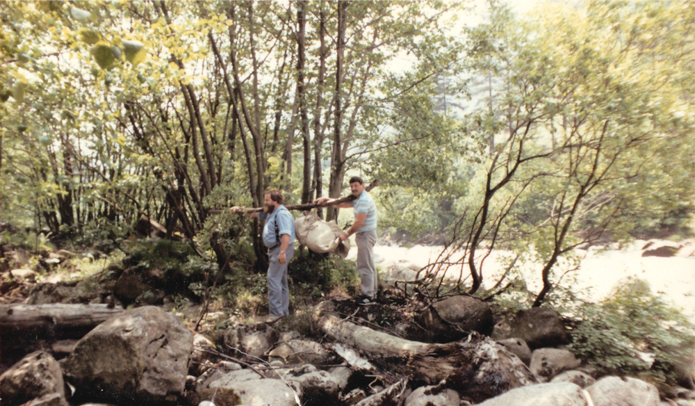 A scan of a photograph from 1985 on a riverbank. Two men carry the Ybrig Stone in a sling hanging from a tree branch across their right shoulders.